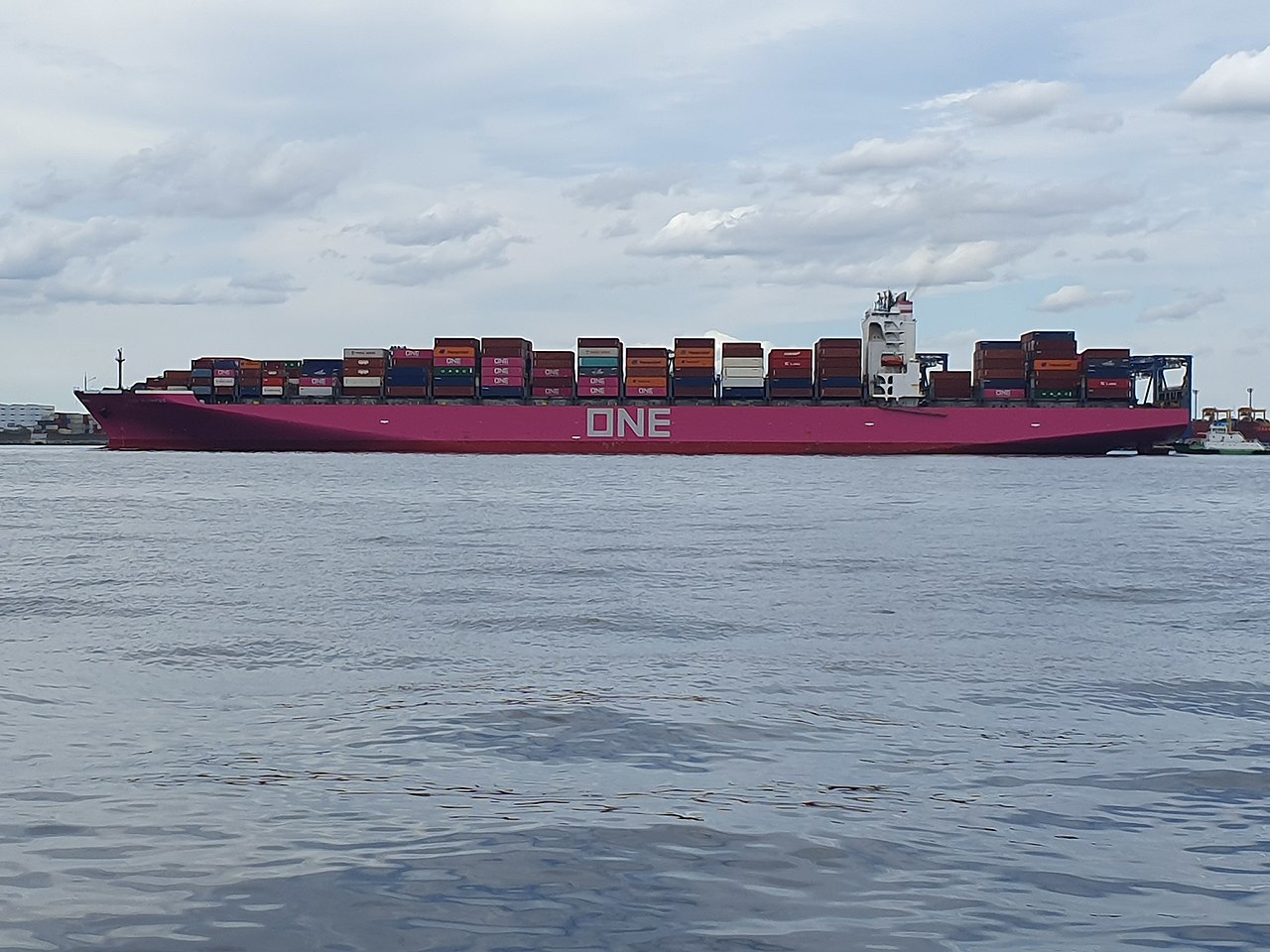 Container ship entering harbor with stacked cargo boxes and port infrastructure.